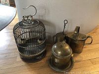 Photo showing brass bird cage with dome top, hook, and brass watering can alongside a planter and fireplace lighter on a wooden floor.