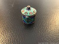 Front view of a small sterling silver jar with intricate turquoise, blue, and green enamel decorative patterns, showing the knob handle on the lid and detailed floral motifs.