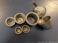 Top-down view showing all six brass nesting measures laid out, with the largest featuring an open hinged lid, highlighting size difference and condition.