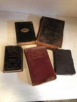 Five antique books displayed on a flat surface, four appear to be Bibles and one is a medical dictionary in red cover.