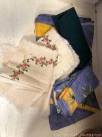 Photo showing folded yellow, blue, and olive green picnic tablecloth, a dark green napkin, and two beige floral embroidered placemats.