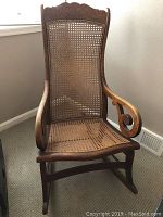 Front view of antique rocking chair showing caned seat and backrest, wooden frame with decorative curved armrests.