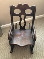 Front view of antique child's rocking chair showing seat, armrests, and backrest with distinctive cutouts.