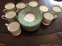 Ten bone china embossed demitasse plates stacked with eight matching cups arranged around them, showing green and gold ornate patterning