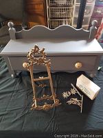 Front view of grey wood wall shelf with drawer, brass decorative holder, pewter hooks, and box of hooks on black fabric.