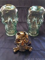 Two large clear glass skulls with hollow interiors and one small metallic skull with crossbones placed in front.