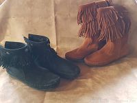 Two pairs of women's Minnetonka moccasins displayed, one black suede pair with fringe and snap closures, one tan leather pair with double fringe layers.