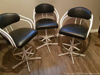 Three black and white bar stools with padded seats and backs, metal frames, footrests, and swivel bases on wood floor.