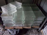 Stacks of clear, ribbed glass blocks arranged on pallets under shelf in storage. Blocks appear square and thick with consistent ribbed texture.