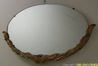Front view of oval antique gilt wood framed mirror hung on wall showing carved ornate design at bottom and sides.