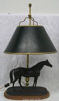 Front view of the antique bronze lamp showing the black lampshade, brass harp, and bronze thoroughbred horse sculpture mounted on an oval base.