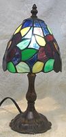 Lamp illuminated and showing stained glass floral shade with varied colored glass pieces and metal base with ornate finish.