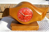 Front view of airplane propeller style wooden clock case with red clock face and white numerals and hands, showing polished wood texture and clock face details.