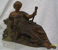 Front and angled view of the Victorian reclining lady metal statue showing draped clothing, holding a staff, and reclined against a decorated back support with floral motifs.