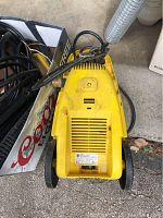 Yellow Karcher K2 electric power washer on a pavement surface with power cord and hose attached. Shows top view with some dirt and wear marks.