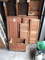 Photo showing assortment of wooden shutters in multiple sizes arranged vertically and stacked inside a garage or storage space. Wooden Coca-Cola crate positioned in front.