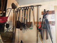 Several long-handled manual garden tools hanging on wall (shovels, spade, fork, rake, hoe).