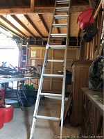 Full length image of the aluminum extension ladder standing in a garage with the ladder extended vertically reaching under the ceiling. The garage contains various tools and storage items.