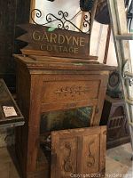 Front and side view of vintage wooden ice chest showing carved floral design and detached front door with scroll carvings, with 'Gardyne Cottage' sign above (not included).