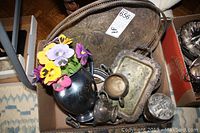 Photo of cardboard box with multiple silver and silver-plated metalware items, including a vase with artificial flowers, a creamer, sugar bowl, and small trays showing tarnish and wear.