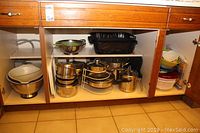 Wide shot of open kitchen cupboard showing pots, pans, and serving trays stacked and arranged inside.
