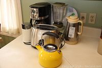 Photo displaying all items on countertop: Oster coffee maker, stainless steel toaster, Sunbeam blender, Braun coffee grinder, yellow kettle, and another white appliance.