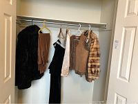 Wide shot of clothing on hangers including black faux fur coat, brown leather vest, beige and black garments, and a fur striped jacket
