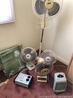 Wide shot showing all fans and heaters included in the lot gathered on the floor near a wall. Includes the tall standing fan, three smaller fans and two heaters as well as the stand fan box.