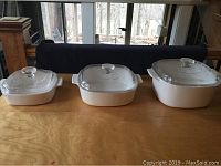 Photo showing three white Corning Ware casserole dishes with glass lids side by side on a wooden surface in front of windows.