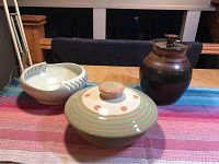 Three signed pottery pieces displayed on a multicolored striped cloth. The pieces include a carved white bowl, a large lidded bowl with striped patterns, and a dark glazed jar with lid.