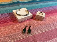 Full set of sterling silver cuff bracelet, plain band ring, and pair of drop earrings with dark oval stones laid out on a multicolored fabric surface.