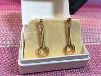 Pair of 14K gold dangle earrings in a cream colored cushioned white box, featuring geometric design with vertical bar and concentric loops.