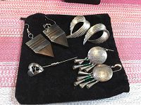 Photo of silver-coloured jewelry items on a black cloth background, includes a long stick pin and three pairs of earrings with different designs.