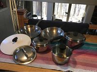 Photo shows seven pots and pans laid out on a table runner, including frying pans, saucepans, and lids, all showing wear from use.