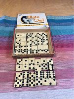 Domino tiles arranged on a multicolored woven textile with the White Owl box lid behind showing the full set of 28 dominoes with ebony pips.
