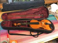 Full view of violin in case alongside bow inside soft black case with red velvet lining.
