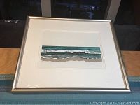 Framed ocean wave artwork in silver frame resting on table with blue woven mat beneath.