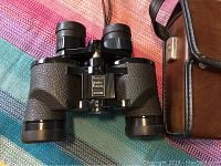 Front view of black textured Bushnell Insta Focus Zoom binoculars and part of the brown carrying case with strap.