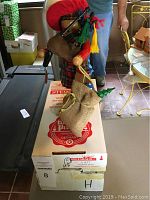 Full side view of Scottish Santa figurine on top of the Steinbach branded box, showing the traditional red hat, plaid pants, bagpipes, and burlap sack with Christmas items.
