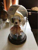Snoopy astronaut figurine inside clear plastic dome on wooden base, shown from front and angled views