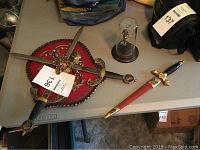 Coat of arms plaque with red background and two crossed swords; letter opener and small plaque with hourglass on table.