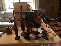 Table display of various carved wooden bird figurines including tall slender forms, ducks, and a small white ceramic dish with bird handles.