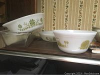 Five vintage Pyrex mixing bowls grouped on a wooden surface, showing green floral and yellow vegetable patterns and a clear rectangular glass baking dish.