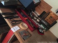 Wide shot showing various hand tools on a wooden surface including scissors, clamps, tape rolls, and a red tool tray with tools inside.