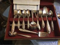 Close-up of silverplate flatware pieces arranged in red velvet-lined case, showing embossed ornate handles and various types of forks and spoons.