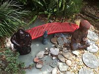 Full view of garden scene with red curved wooden bridge, two Buddha statues in dark finish sitting on stones and soil with greenery surrounding