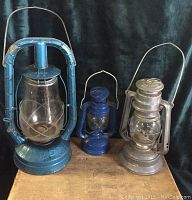 Front view of three vintage lanterns: large blue, small blue, and medium gray lanterns on wooden surface against dark background.
