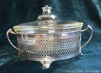 Side view of oval Pyrex glass casserole in ornate silver plate frame with handles and feet, with glass lid on top.