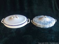 Two oval covered porcelain bowls displayed side by side on a dark background. One bowl features a simple blue line detail and double loop handle on the lid, the other has blue floral patterns and a sculpted leaf and bird knob.