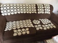 All nine crocheted white or cream-colored items displayed on a brown textured couch, including runners and placemats of varying sizes and shapes, highlighting the floral motifs and overall condition.